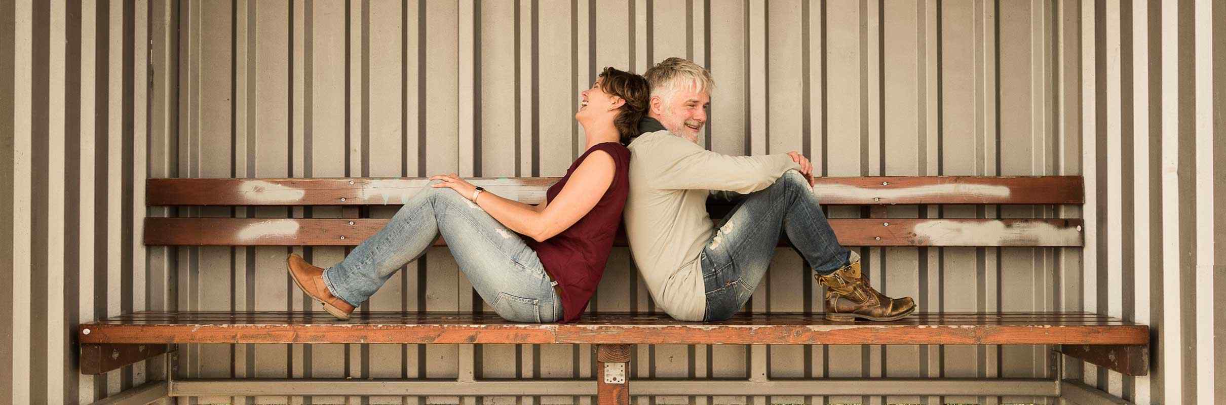 Paar sitzt lachend Rücken an Rücken auf Bank an Schiffshaltestelle, Norddeutsche Parakademie, Ausbildung in Paartherapie